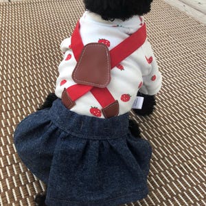 May include: A white long-sleeve shirt with a red strawberry print paired with a dark blue denim skirt. The shirt features red elastic braces with brown faux leather accents that cross over the back, creating a layered outfit for a small pet.
