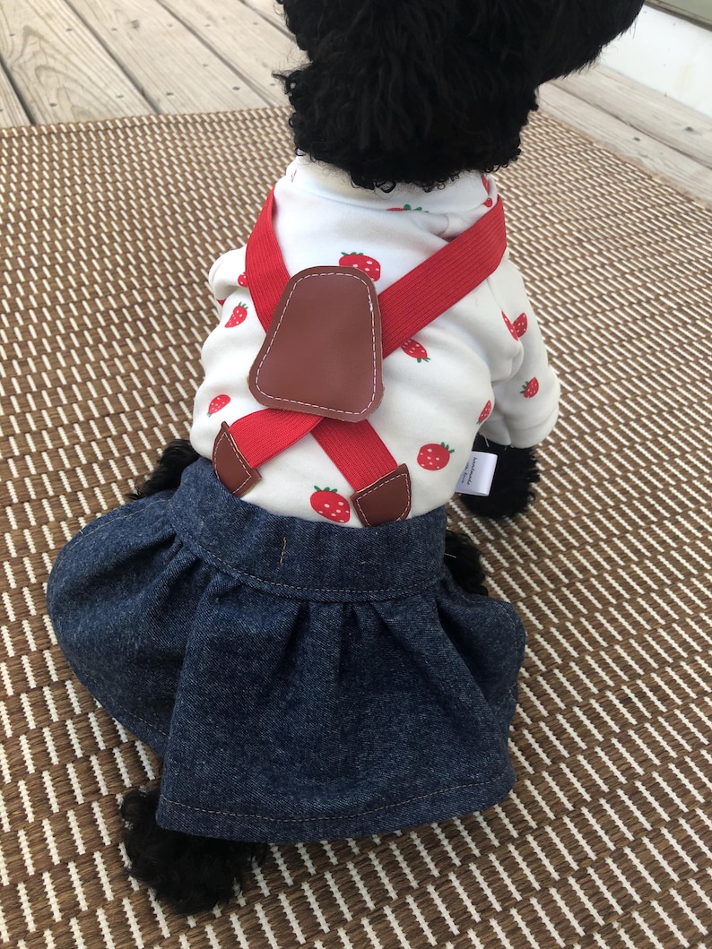 May include: A white long-sleeve shirt with a red strawberry print paired with a dark blue denim skirt. The shirt features red elastic braces with brown faux leather accents that cross over the back, creating a layered outfit for a small pet.