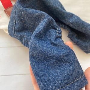 May include: A pair of blue denim dungarees featuring a white bib with a red strawberry print and bright red adjustable straps. The denim fabric has a textured appearance with visible stitching along the hem and gathered elastic sides for a flexible fit.