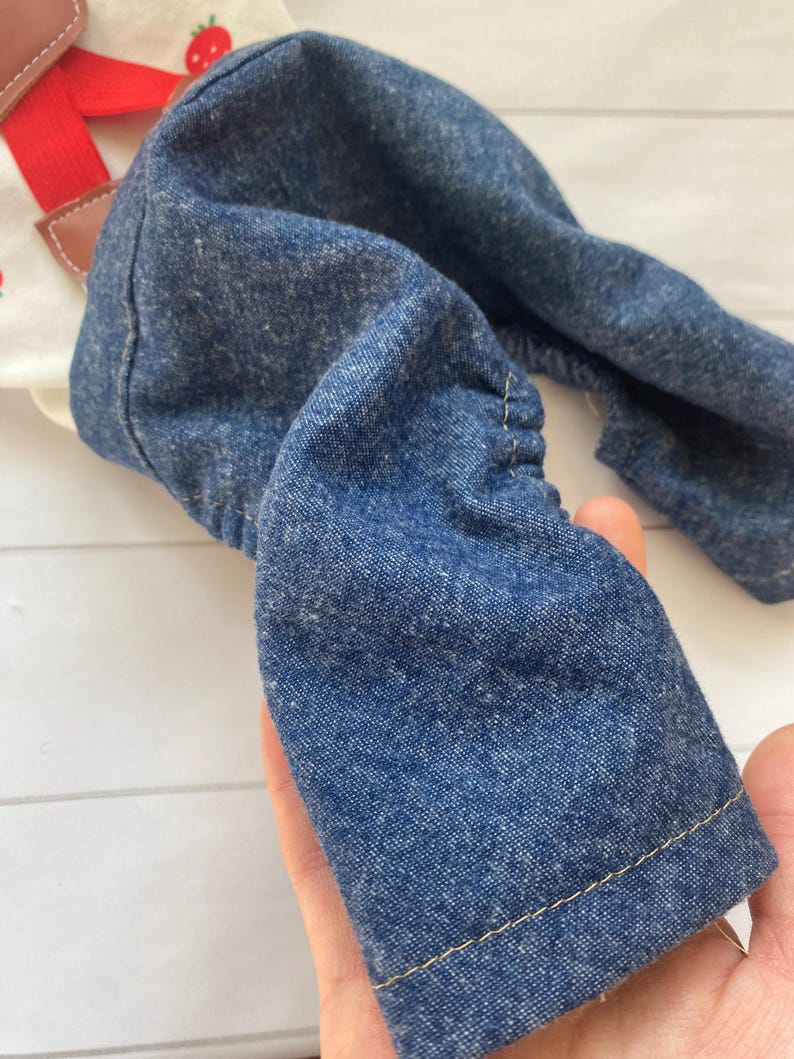 May include: A pair of blue denim dungarees featuring a white bib with a red strawberry print and bright red adjustable straps. The denim fabric has a textured appearance with visible stitching along the hem and gathered elastic sides for a flexible fit.