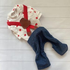 May include: A white short-sleeved shirt featuring a red strawberry print, paired with red elastic suspenders that have brown faux leather accents. The outfit includes dark blue denim trousers with an elasticated waist and leg openings.
