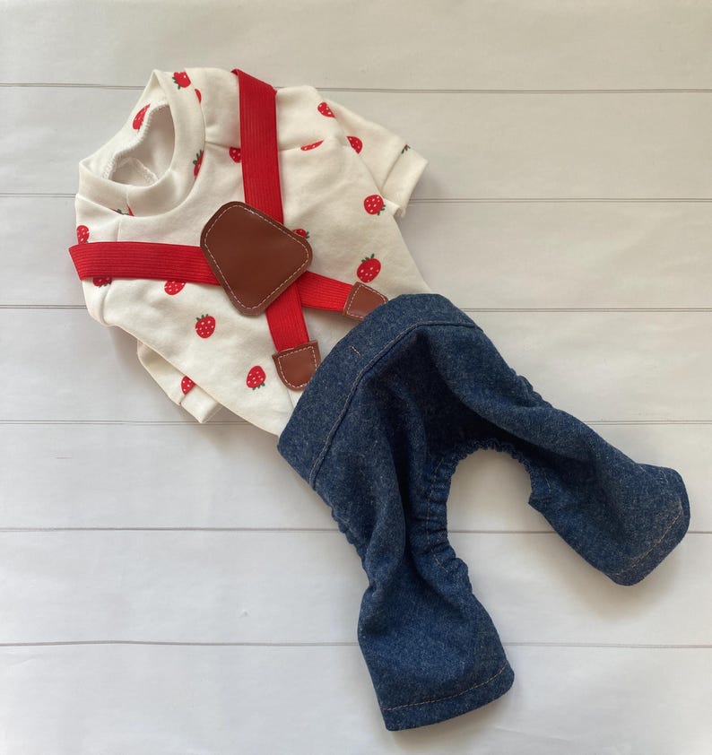 May include: A white short-sleeved shirt featuring a red strawberry print, paired with red elastic suspenders that have brown faux leather accents. The outfit includes dark blue denim trousers with an elasticated waist and leg openings.
