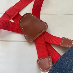 May include: Bright red elastic braces featuring a brown faux leather Y-back patch and matching brown faux leather tabs with white stitching. The braces are attached to a piece of blue denim fabric at the bottom edge of the frame.