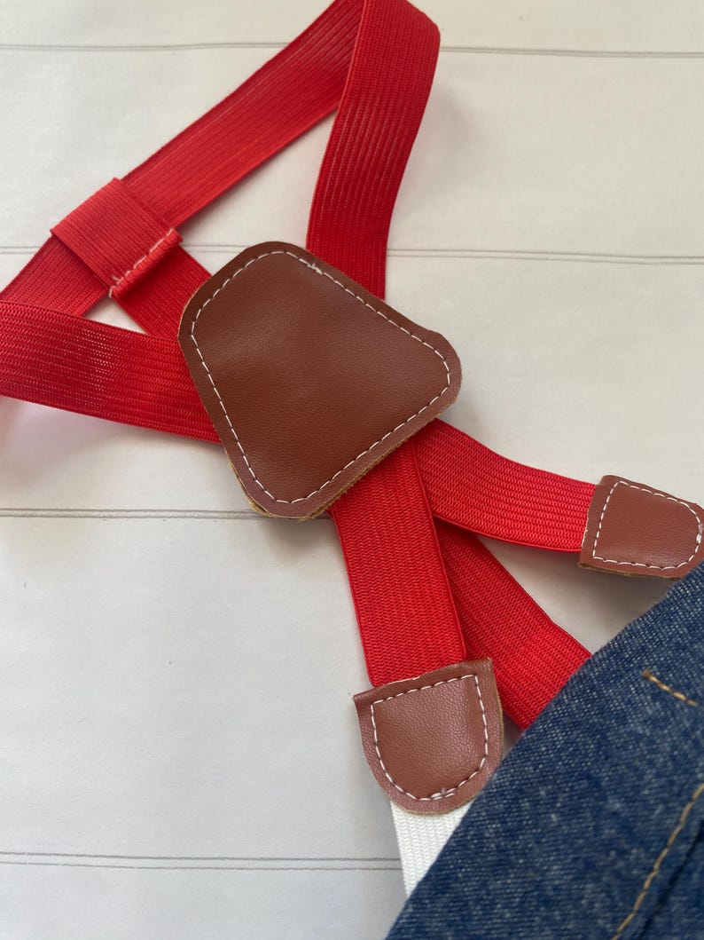 May include: Bright red elastic braces featuring a brown faux leather Y-back patch and matching brown faux leather tabs with white stitching. The braces are attached to a piece of blue denim fabric at the bottom edge of the frame.
