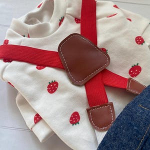 May include: A white long-sleeved shirt featuring a red strawberry pattern, paired with red elastic braces that have brown faux leather accents. The braces feature a central brown faux leather patch where the straps cross over the chest.