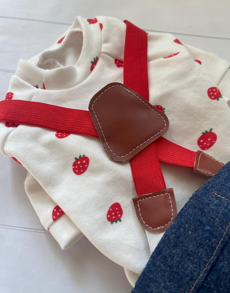 May include: A white long-sleeved shirt featuring a red strawberry pattern, paired with red elastic braces that have brown faux leather accents. The braces feature a central brown faux leather patch where the straps cross over the chest.
