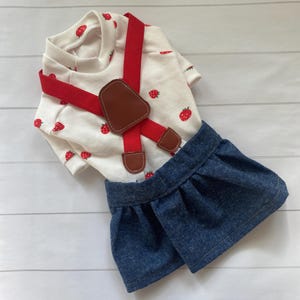 May include: A white short-sleeved shirt featuring a red strawberry pattern paired with a dark blue denim skirt. The outfit includes bright red elastic braces with brown faux leather accents at the attachment points and the centre front.