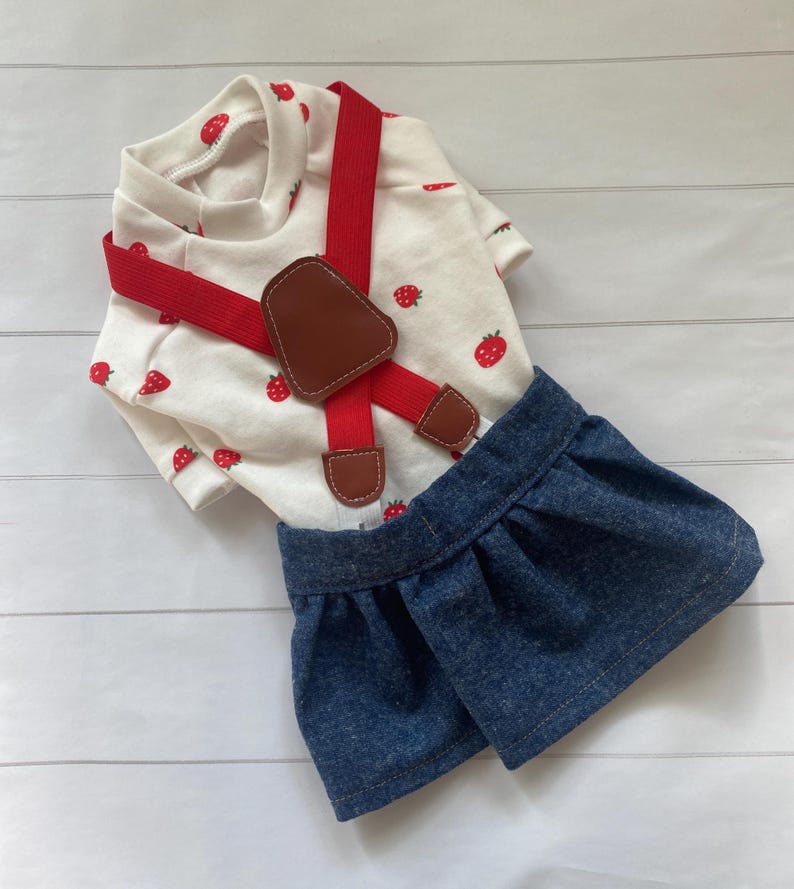 May include: A white short-sleeved shirt featuring a red strawberry pattern paired with a dark blue denim skirt. The outfit includes bright red elastic braces with brown faux leather accents at the attachment points and the centre front.