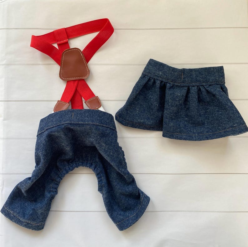 May include: A dark blue denim overall set featuring red elastic braces with brown leather-look accents and a matching dark blue denim pleated skirt. The overalls have a gathered waist and leg openings, while the skirt has a simple waistband.