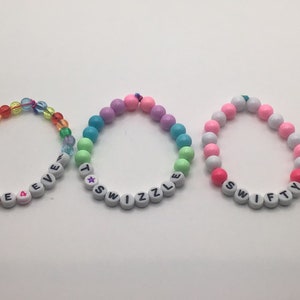 Puede incluir: Tres pulseras de cuentas de colores con cuentas de letras blancas que deletrean las palabras "SWIFTIE", "TWIZZLE" y "SWIFTY".