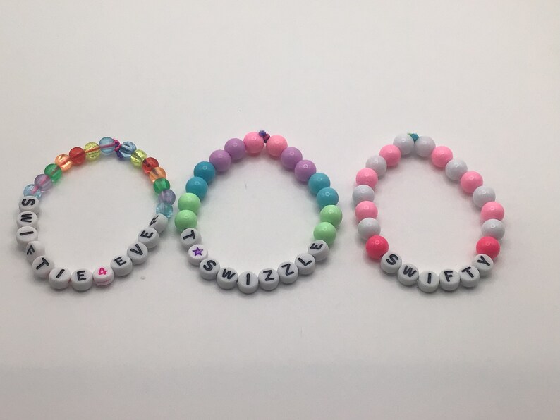 Puede incluir: Tres pulseras de cuentas de colores con cuentas de letras blancas que deletrean las palabras "SWIFTIE", "TWIZZLE" y "SWIFTY".