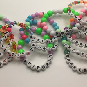 Puede incluir: Una colecci&oacute;n de pulseras de cuentas coloridas con diferentes nombres escritos en ellas, incluyendo "Olivia", "Best", "Last", "Sparks", "Speak", "Know", "Epiphany", "August", "Wizzl", "Era", "Fifty", "Chance", "Tergl" y "Midnights".