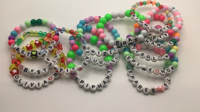 Puede incluir: Una colecci&oacute;n de pulseras de cuentas coloridas con diferentes nombres escritos en ellas, incluyendo "Olivia", "Best", "Last", "Sparks", "Speak", "Know", "Epiphany", "August", "Wizzl", "Era", "Fifty", "Chance", "Tergl" y "Midnights".