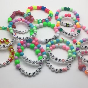 Puede incluir: Una colecci&oacute;n de pulseras de cuentas de colores con varias palabras escritas, incluyendo "Best", "Olivia", "Fearless", "Swizzle", "Eras", "Change", "Speak Now", "August", "Epiphany", "Sparks", "Fifty", "Afterglow", "Karma", "Bad Blood" y "Stick".