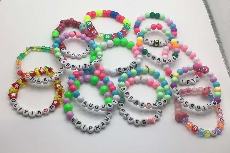 Puede incluir: Una colecci&oacute;n de pulseras de cuentas de colores con varias palabras escritas, incluyendo "Best", "Olivia", "Fearless", "Swizzle", "Eras", "Change", "Speak Now", "August", "Epiphany", "Sparks", "Fifty", "Afterglow", "Karma", "Bad Blood" y "Stick".
