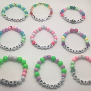 Puede incluir: Nueve pulseras de cuentas de colores con diferentes palabras escritas en ellas. Las palabras son "Speak Now", "Best Friends", "Last Kiss", "T*Swizzle", "Swifty", "Afterglow", "Eras", "Epiphany" y "Sparks Fly".