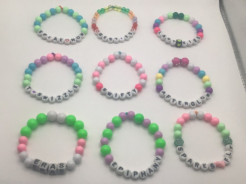 Puede incluir: Nueve pulseras de cuentas de colores con diferentes palabras escritas en ellas. Las palabras son "Speak Now", "Best Friends", "Last Kiss", "T*Swizzle", "Swifty", "Afterglow", "Eras", "Epiphany" y "Sparks Fly".