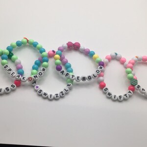 Puede incluir: Cinco pulseras de cuentas de colores con diferentes palabras escritas en ellas. Las pulseras son: "Last Kiss", "Speak Now", "Afterglow", "Swifty" y "Sparks Fly".