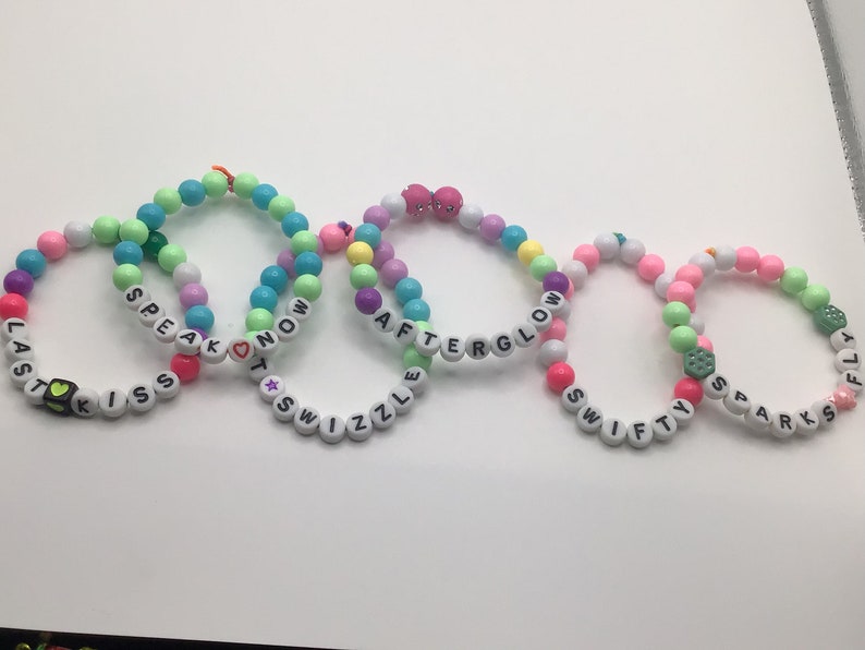 Puede incluir: Cinco pulseras de cuentas de colores con diferentes palabras escritas en ellas. Las pulseras son: "Last Kiss", "Speak Now", "Afterglow", "Swifty" y "Sparks Fly".