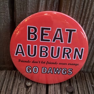 May include: Red round button with the text "BEAT AUBURN" in black letters. Below the text is the phrase "Friends don't let friends wear orange" in smaller black letters. The bottom of the button says "GO DAWGS" in black letters.