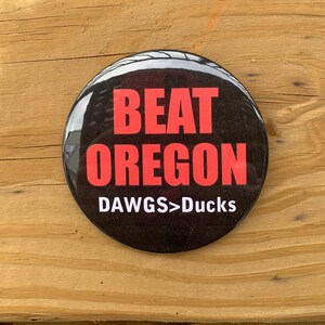 May include: A black button with the text "BEAT OREGON" in red letters and "DAWGS>Ducks" in white letters.