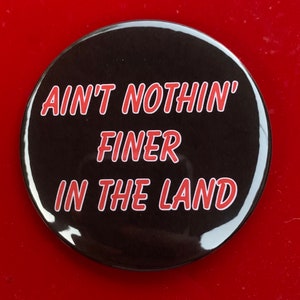 Puede incluir: Un botón negro con texto blanco que dice "Ain't nothin' finer in the land" en rojo.
