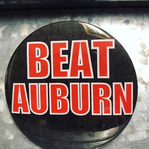 Puede incluir: Un botón negro con borde blanco y las palabras "BEAT AUBURN" en rojo.