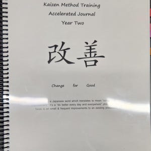 Può includere: Un diario bianco con una rilegatura a spirale nera. La copertina presenta il testo "Kaizen Method Training Accelerated Journal Year Two" e i caratteri giapponesi "改善" che si traducono in "Change for Good".