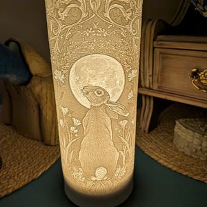 Può includere: Lampada cilindrica bianco sporco con un design a rilievo dettagliato. Il design presenta un coniglio che guarda una luna piena, circondato da elementi floreali e fogliari. La lampada è illuminata dall'interno, creando una luce soffusa.