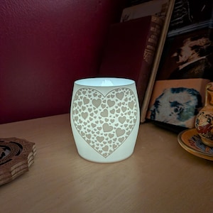 May include: A glowing white candle holder with a heart-shaped design filled with smaller hearts. The candle holder is cylindrical and sits on a wooden surface. The background includes books and a dark red wall.