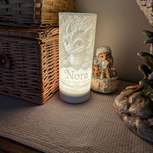 May include: A cylindrical white night light featuring a 3D-printed relief of a baby dragon. The front surface displays the name Nora and the date 6th March 2026 in a decorative font. The lamp emits a soft glow from within its textured structure.