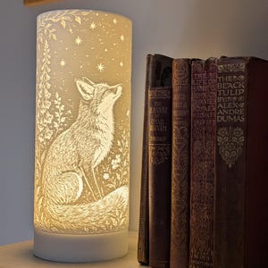May include: A cylindrical white lamp featuring an illuminated relief design of a fox sitting among flowers and stars. The fox is looking upward toward the stars. The lamp is positioned next to a row of four antique books with visible text.