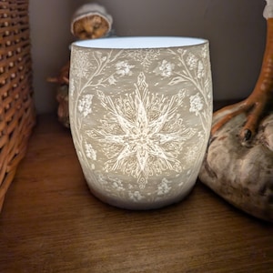 May include: An illuminated, off-white glass candle holder featuring a snowflake and floral design. The cylindrical holder is lit from within, highlighting the intricate patterns. The candle holder is approximately 10 cm tall.