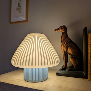 May include: A decorative table lamp with a mushroom-shaped lampshade and a light blue base. The lampshade is pleated and off-white. A bronze dog statue and framed artwork are on a wooden surface. The lamp is lit, providing a warm light.