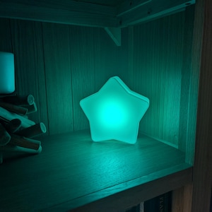 May include: A white star-shaped nightlight with a teal glow, sitting on a wooden shelf.