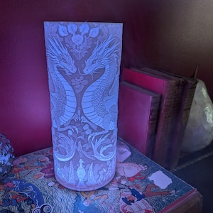 May include: A white, illuminated cylindrical lamp featuring a detailed dragon design. The lamp displays two dragons facing each other amidst floral patterns. It sits on a table with books and a decorative glass jar.