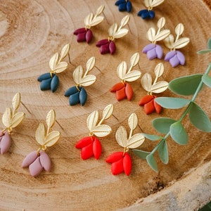 May include: A collection of stud earrings featuring gold-toned metal leaves attached to small, matte-finished flower petals in various colors including navy, burgundy, lavender, teal, orange, red, and dusty pink, arranged in pairs on a wooden surface.