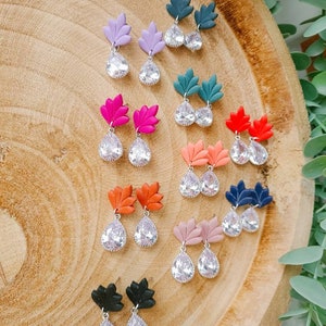 Puede incluir: Una colección de nueve pares de pendientes, cada uno con un componente superior en forma de hoja con acabado mate en varios colores como lavanda, verde azulado, rosa, coral, naranja, rosa viejo, azul marino y negro, junto a un cristal de lágrima.