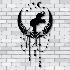 Puede incluir: Arte de pared de metal negro con la silueta de un elefante sobre una luna creciente, con estrellas y cuentas colgantes. La luna tiene un patrón detallado. La obra de arte está montada en una pared de ladrillos blancos.