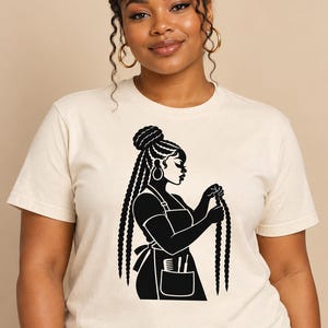 Puede incluir: Una camiseta de manga corta de color crema con un estampado gráfico negro de una persona con el pelo largo trenzado que lleva un delantal. El gráfico muestra a la persona sosteniendo una trenza larga, con un peine y tijeras en el bolsillo.