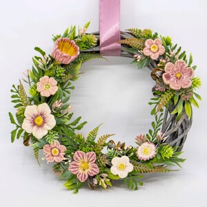 May include: A pink and green floral wreath with a pink ribbon hanging from the top. The wreath is made of woven twigs and has a variety of pink flowers and green leaves.