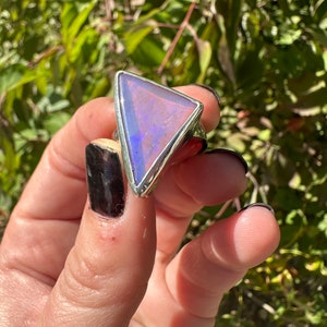 May include: A silver ring with a triangular, iridescent opal stone. The opal displays shades of pink, purple, and blue. The ring is set against a backdrop of green foliage, highlighting the gemstone's colors.