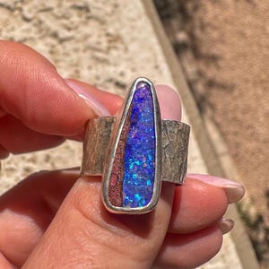 May include: A silver ring featuring a teardrop-shaped inlay of iridescent blue and purple opal alongside a textured reddish-brown stone segment. The band of the ring has a hammered, metallic finish with a wide, flat profile and polished edges.