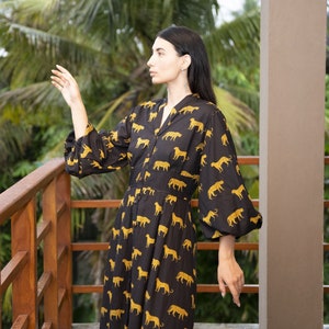 May include: A woman wearing a black and gold long-sleeved dress with a leopard print pattern. She is standing on a wooden balcony with a lush green background.