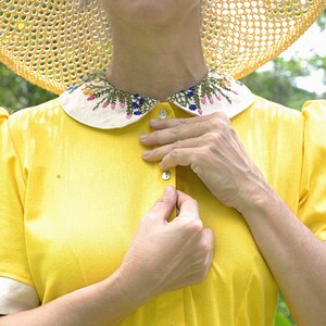 May include: A woman wearing a yellow dress with a white collar embroidered with flowers. She is fastening a button on the front of the dress.