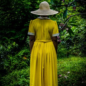 May include: A woman wearing a yellow dress and a wide-brimmed straw hat stands in a lush green garden.