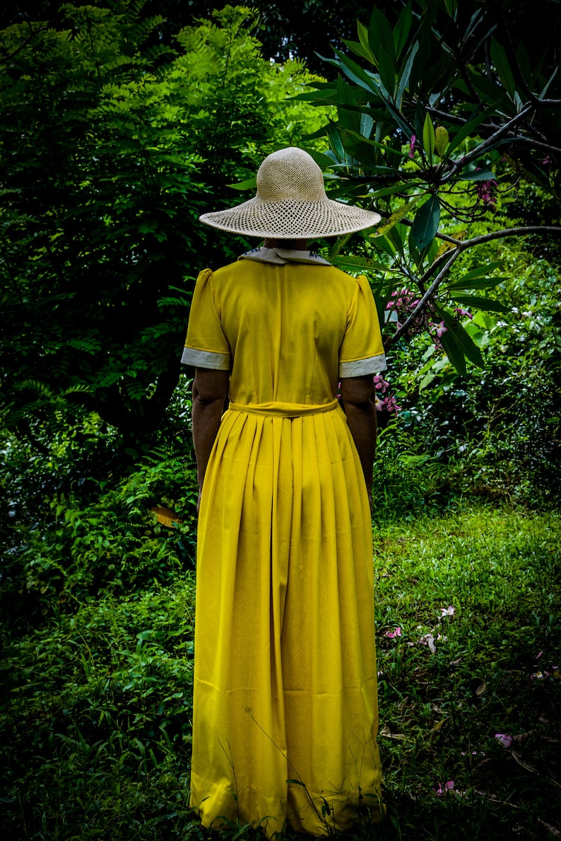 May include: A woman wearing a yellow dress and a wide-brimmed straw hat stands in a lush green garden.