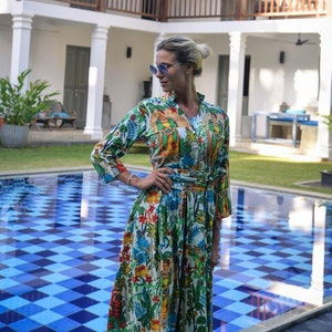 May include: A woman wearing a white maxi dress with a colorful floral print and a tropical theme. She is standing by a blue and white checkered tiled pool.
