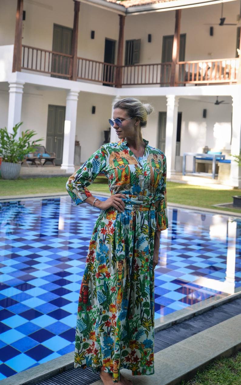 May include: A woman wearing a white maxi dress with a colorful floral print and a tropical theme. She is standing by a blue and white checkered tiled pool.