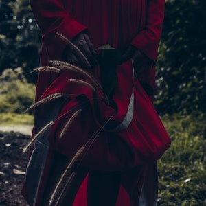 May include: A person wearing a long, red dress with a white belt. The dress is flowing and has a loose fit. The person is standing in a forest setting with tall grass and trees in the background.